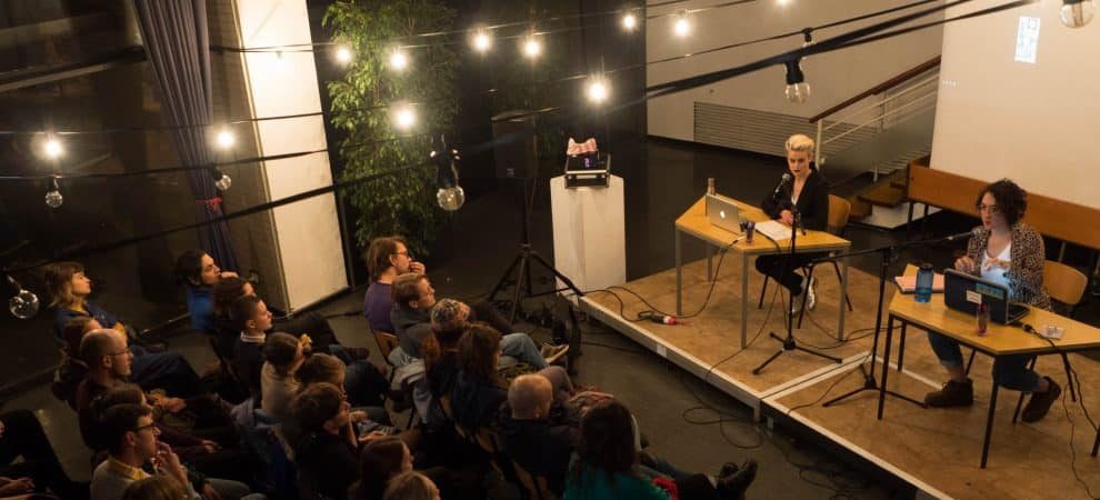 DE: zwei Menschen sitzen auf einer Bühne an Tischen vor ihren Laptops. Zuschauendenreihen davor//EN: two people sitting on a stage at tables infront of their laptops. Rows of spectators in front